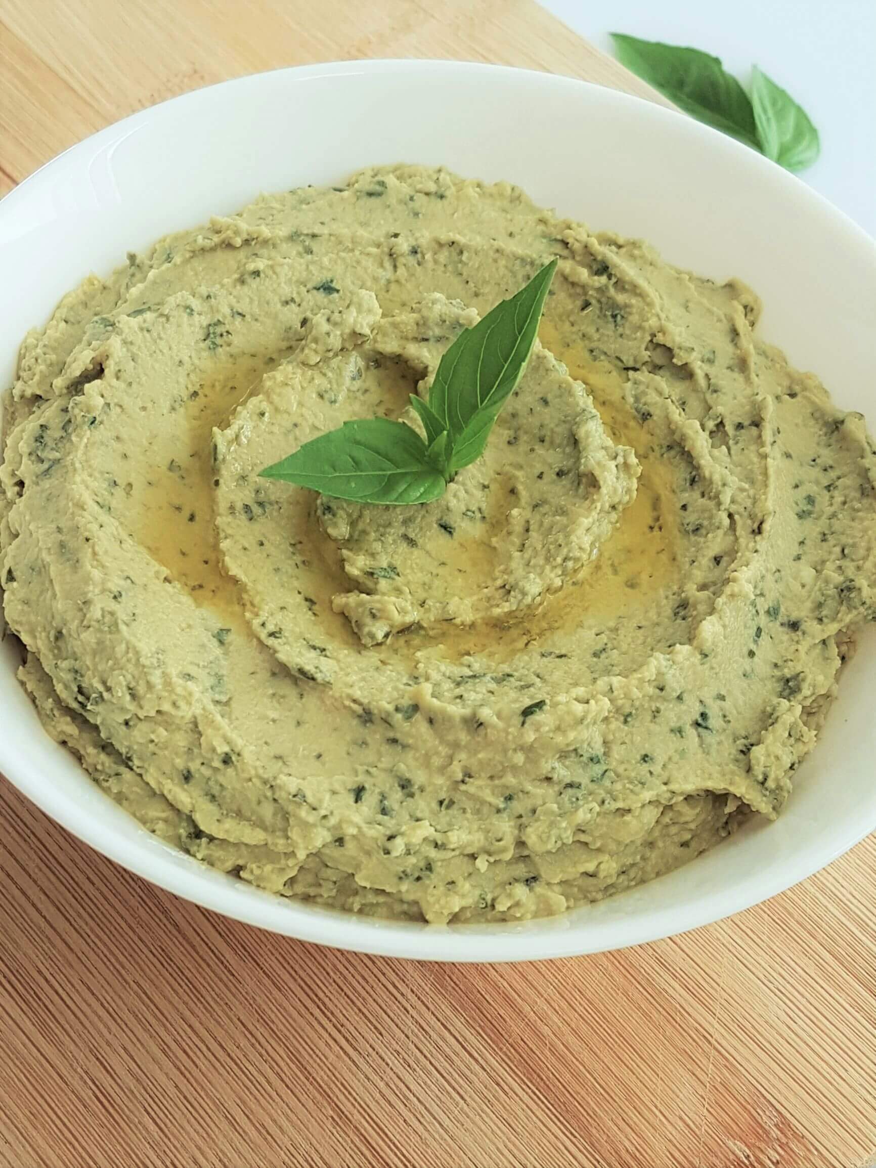 A plate of green basil hummus garnished with a pick of basil leaves in the center and drizzled with olive oil