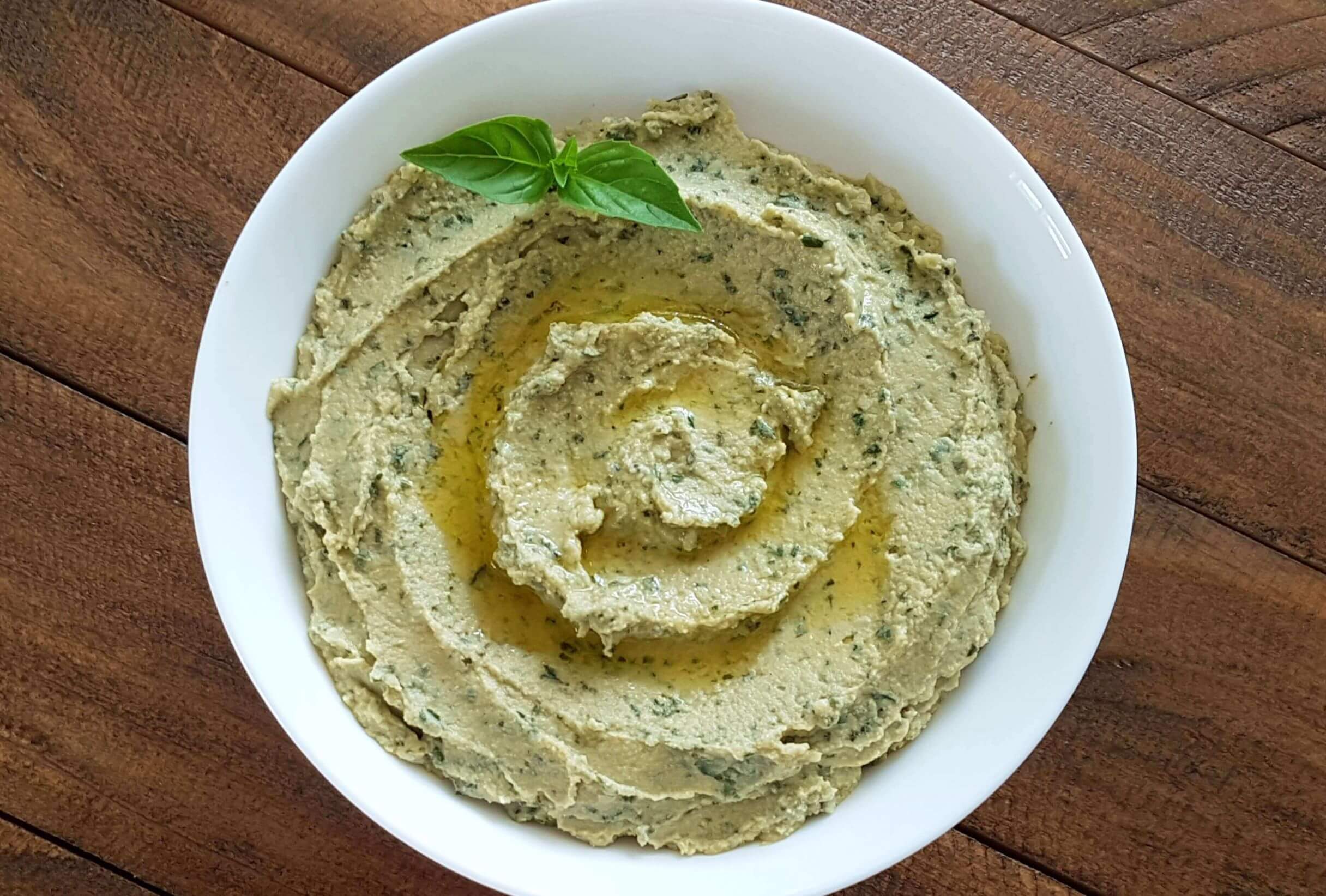 A plate of green basil hummus drizzled with olive oil, and garnished with basil leaves on the edge of the plate.