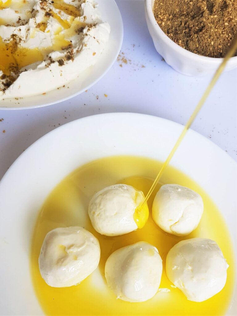 A white plate with five balls of labneh (yogurt cheese) in oil with olive oil being poured on them. In the background, a saucer with Zaatar, and the edge of a plate with Labneh, olive oil and Zaatar.